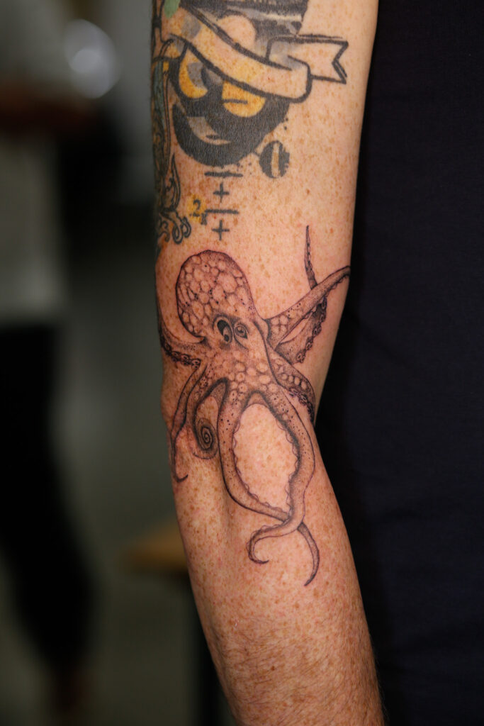 A fineline black and grey octopus tattoo on a freckled forearm. The design shows the octopus with its tentacles gracefully extended.