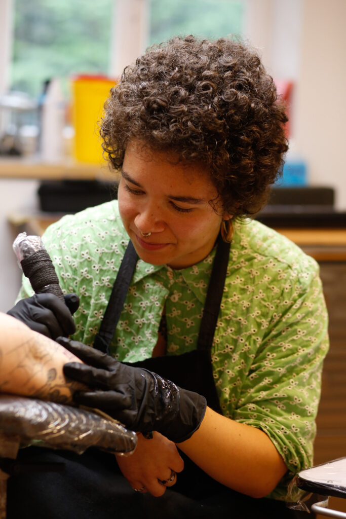 A portrait of tattoo artist Fides from Azramati tattoo with curly dark hair and a septum piercing. She is wearing a green patterned shirt and a black apron, focused on applying a tattoo to a client’s arm.