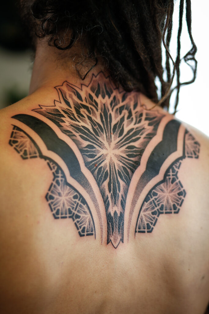 Een grote, symmetrische zwarte tatoeage op de bovenrug en nek met een gedurfde mandala en complexe geometrische lijnen in dotwork, gezet door Bence van BenHTattoo.