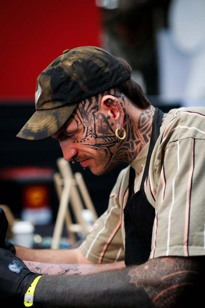 Een portret van tatoeëerder Bence van BenHTattoo, die een tie-dye pet draagt en uitgebreide ornamentale tatoeages op zijn gezicht, nek en armen heeft.