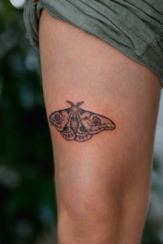A small, intricately detailed black and grey moth tattoo with spread wings, positioned on a person’s thigh.