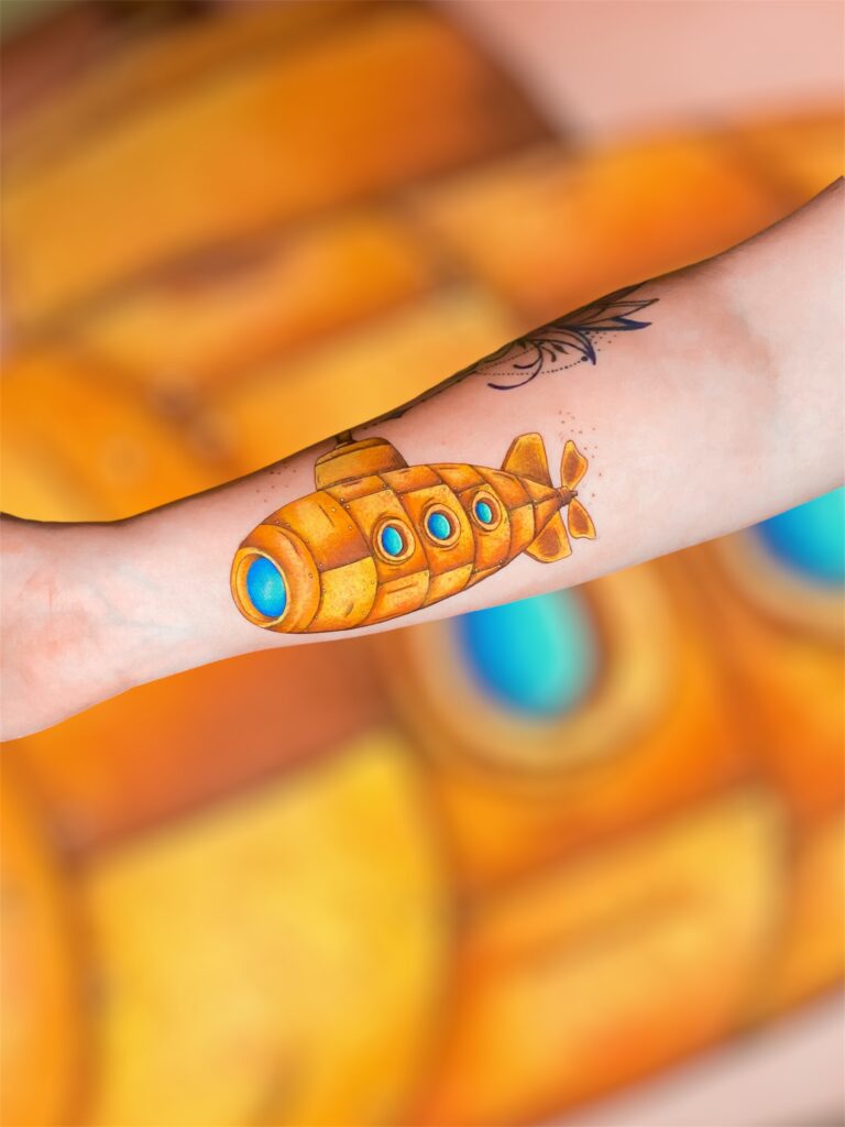 A bright, pop-art inspired yellow submarine tattoo with blue portholes on a forearm, created by Lavinja of LesDix Noirs.