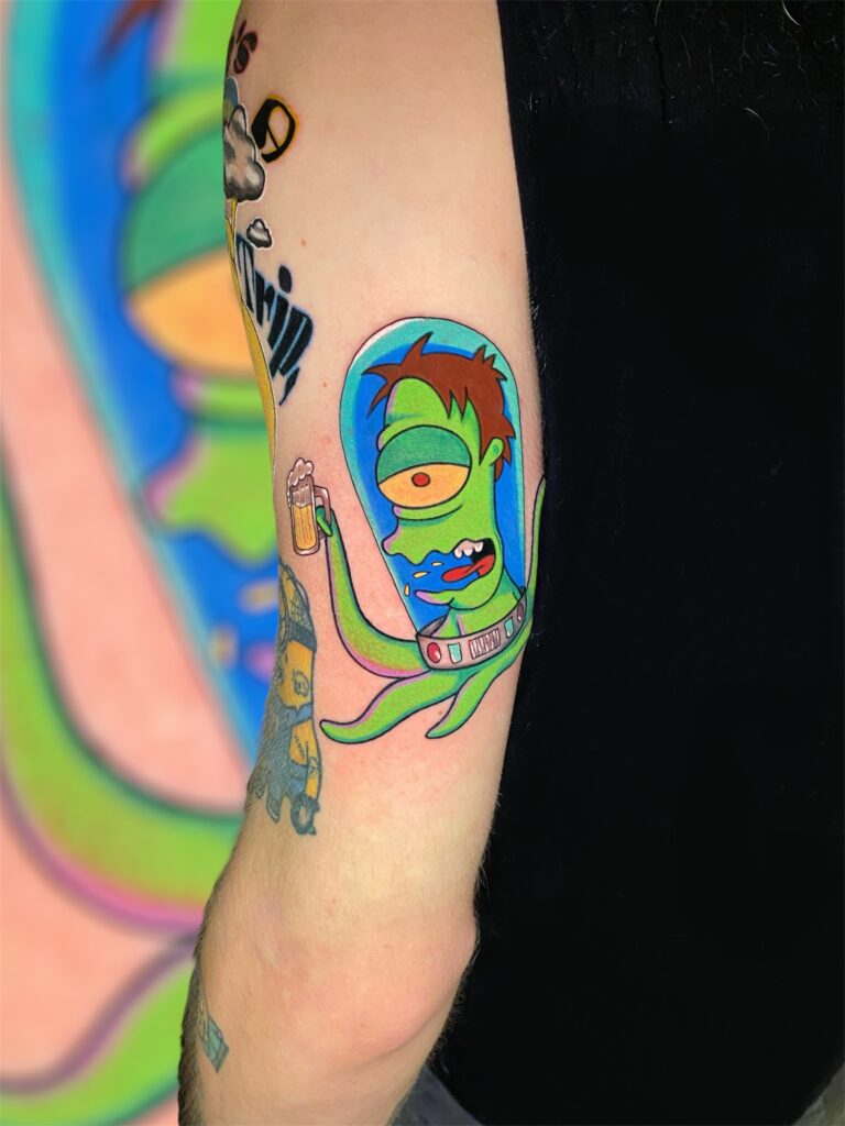 A vibrant, cartoon-style tattoo of a one-eyed alien in a space helmet holding a beer, custom-designed and tattooed by Lavinja of LesDix Noirs.