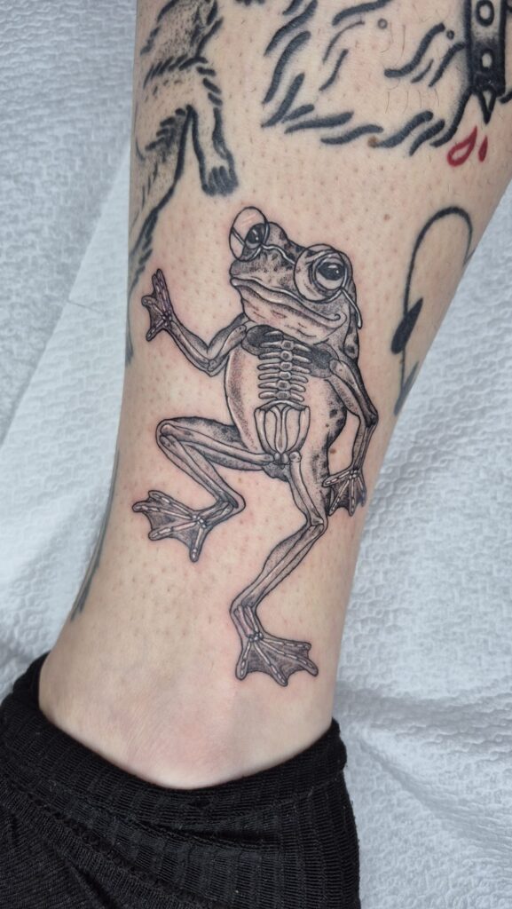 A whimsical black and grey tattoo by Najate from Empowering Life on a leg. It depicts a frog wearing glasses, with its internal skeletal system visible as if viewed through an x-ray.