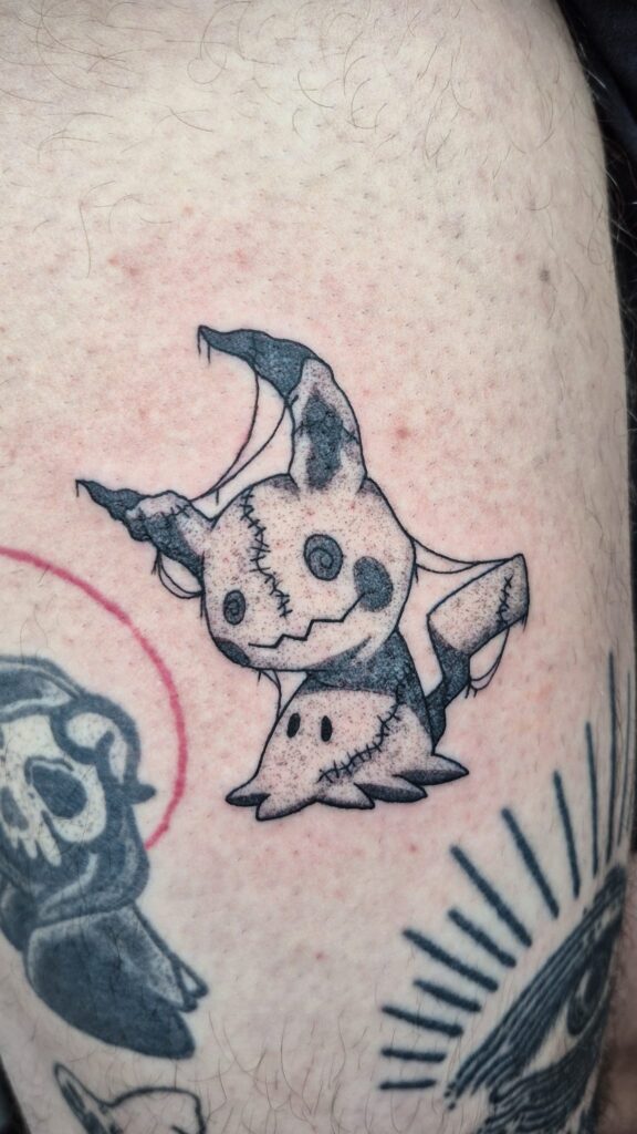 A stylized black and grey tattoo of the Pokémon Mimikyu on a person's leg. Created by Najate of Empowering Life, the design features a gritty, hand-sketched aesthetic with delicate linework and dotwork shading.