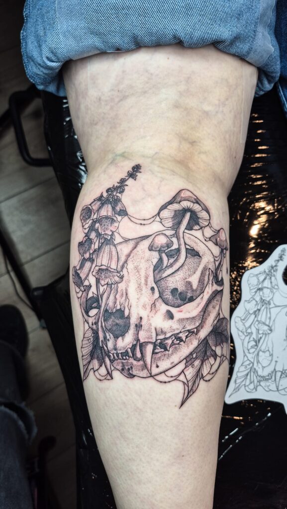 A detailed black and grey tattoo on a limb by Najate of Empowering Life. The piece features a feline skull with mushrooms and foxglove flowers growing out of and around it, rendered with fine dotwork and shading.