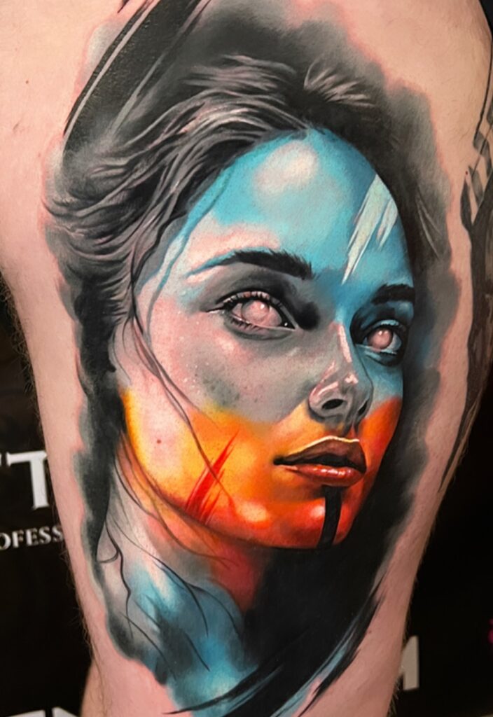 A stunning, colorful portrait tattoo of a woman's face on a client's skin, created by Kenny of Hannon Tattoo. The design features realistic facial features blended with bold, painterly splashes of blue, orange, and red ink.