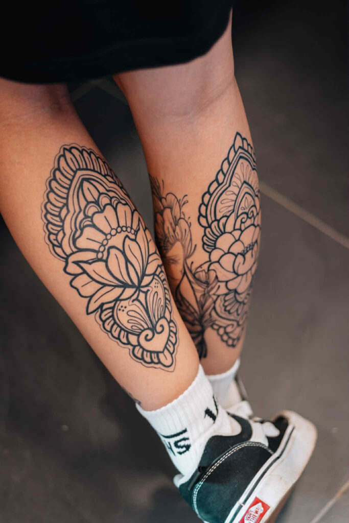 A pair of large, symmetrical ornamental tattoos on a client's shins by Celia of Poppy Lia. The designs feature bold linework, lotus-inspired floral elements, and intricate mandala patterns.