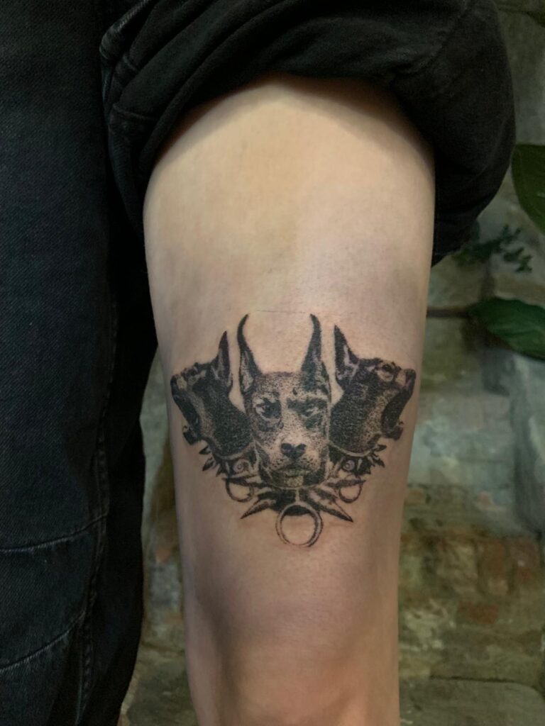 A black and grey tattoo on a thigh depicting three Doberman pinscher heads. The center dog faces forward while the two side dogs are shown in profile, featuring fine dot-work shading and spiked collar details.