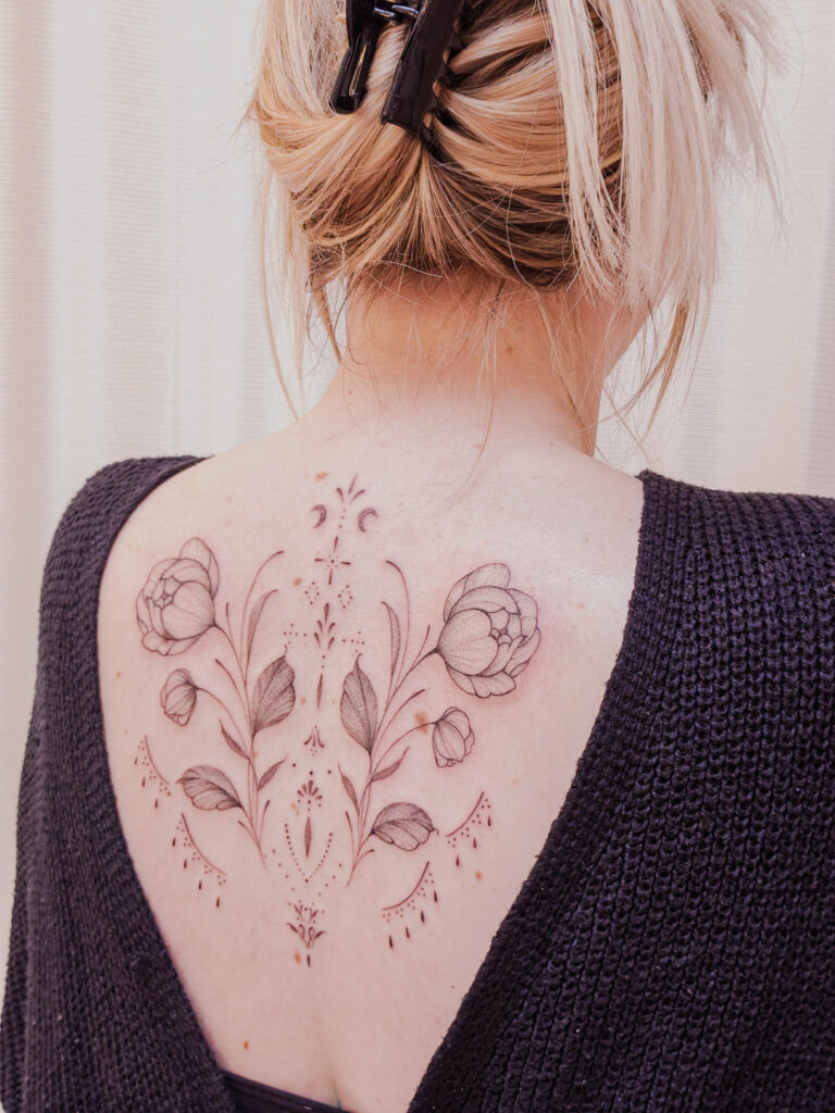 A large, symmetrical fine-line tattoo on a client's upper back by Tacha of Studio Manoja. The piece features two blooming peonies surrounded by intricate ornamental linework and delicate botanical patterns.