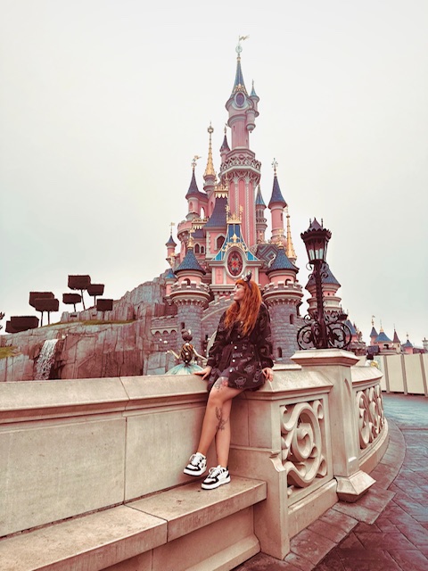 A portrait of tattoo artist Amber from Fantasy Ink. She has long, vibrant red hair and is pictured sitting on a stone wall with the iconic pink Sleeping Beauty Castle at Disneyland Paris in the background.