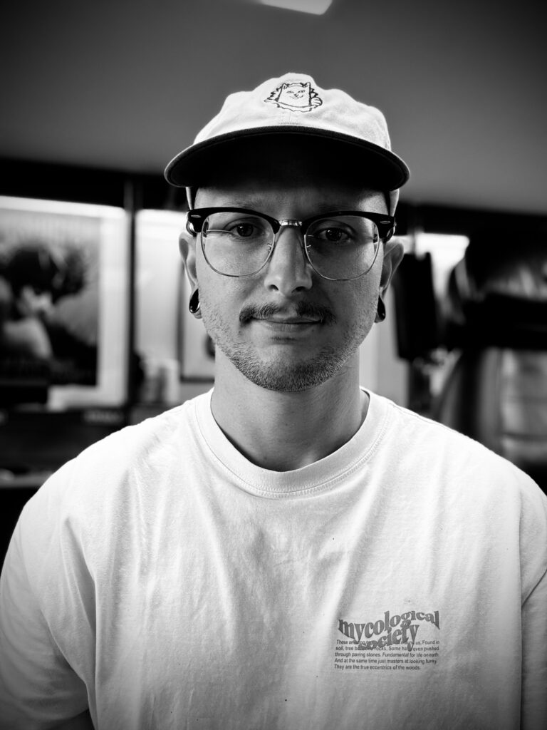 A black and white portrait of tattoo artist Sabbe from T-paint/Roxy. He is wearing glasses, a baseball cap with a small cat logo, and a white t-shirt featuring "mycological society" text. He also has stretched earlobes.