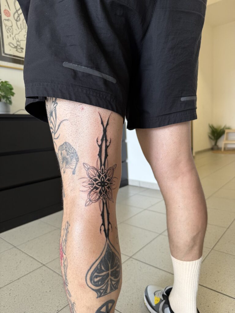 A bold, black-ink tattoo on a thigh by Britt of Inked by Britt. The design features a vertical, sharp "cyber-sigilism" style ornament centered around a geometric star-like mandala.