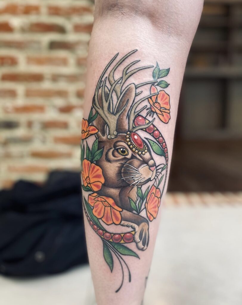 A vibrant, illustrative color tattoo on a leg by Christa of Obscuria Gratia. It features a brown rabbit with antlers (a jackalope) wearing an ornate red jeweled headpiece, surrounded by bright orange poppies and green leaves.