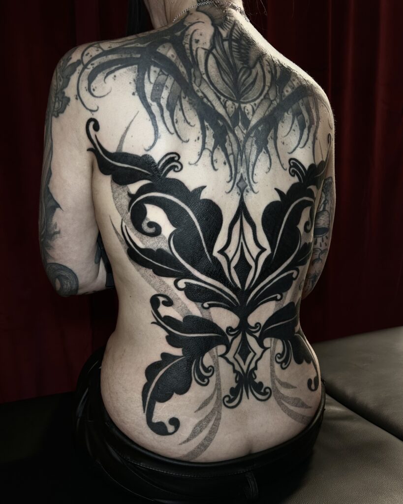 A massive and striking blackwork tattoo covering a person's entire back, created by Helene from Donkerwerk. The piece features thick, flowing ornamental shapes and sharp, symmetrical flourishes that create a powerful, dark silhouette.