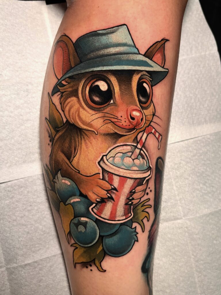A vibrant, illustrative tattoo on an arm by Sabbe of T-paint/Roxy. It features a cute, wide-eyed cartoon possum wearing a blue fedora, holding a red-and-white striped cup with a blue drink and a straw, surrounded by blue berries.