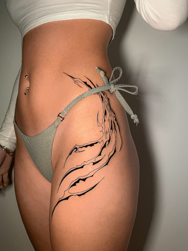 A flowing, black-ink abstract tattoo on a client's hip and upper thigh, created by Britt from Inked by Britt. The design features sharp, flame-like or claw-like curves that follow the natural contour of the body.