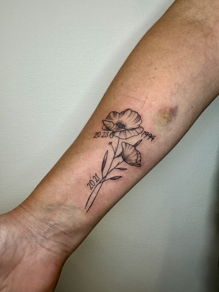 A delicate black and grey floral tattoo on an inner forearm by Laura of Inked By Laura. The design features two blooming flowers with the meaningful years "2023", "1994", and "2021" elegantly scripted around the stems.