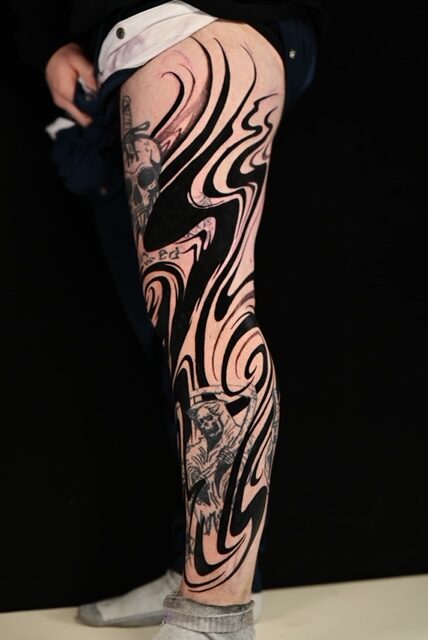 A bold, abstract blackwork tattoo on the back of a leg, created by Ezra of TattooGast. The piece consists of large, flowing black shapes with a liquid-like appearance that extends from the upper thigh down to the ankle.