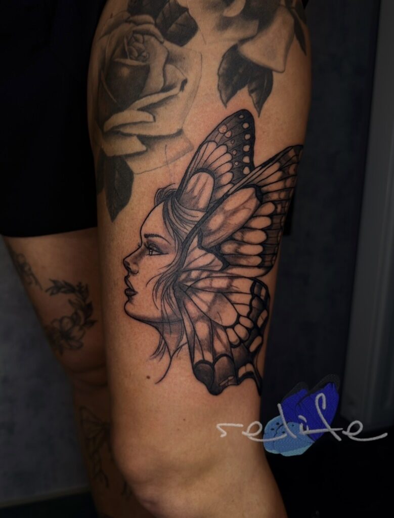 A detailed black and grey tattoo on a leg by Remi from Relife Tattoo. It depicts the side profile of a woman whose head seamlessly merges into the large, intricate wing of a butterfly, set against a background of soft-shaded roses.