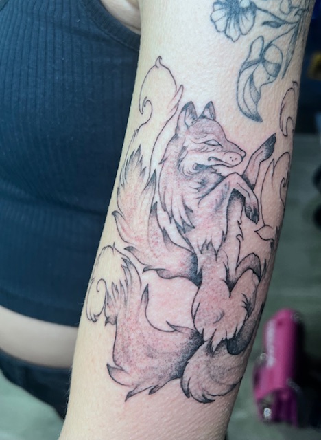 A fineline black and grey tattoo on an arm depicting a mystical multi-tailed fox (Kitsune) in a graceful, flowing pose, created by Bieke of Bibi Ink.