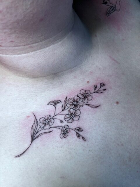 A delicate and precise fineline tattoo of a flowering branch with small, soft blossoms, positioned on a person's upper chest near the collarbone, by artist Bieke from Bibi Ink.