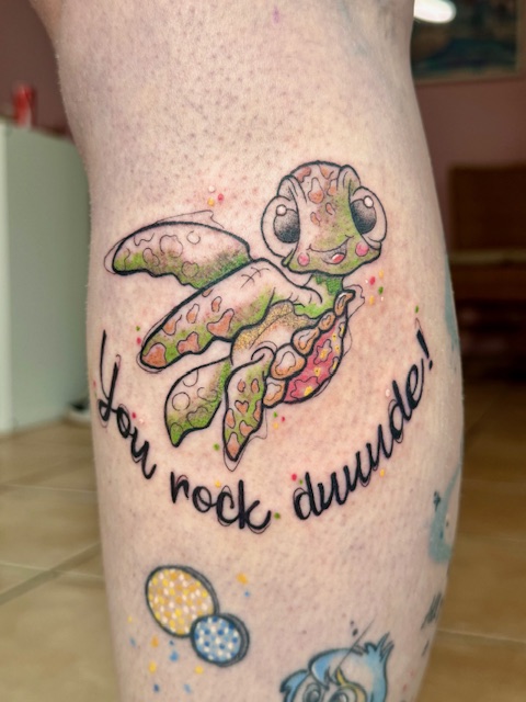 A bright and colorful tattoo on a leg featuring the character Squirtle from Finding Nemo. The turtle is surrounded by colorful bubbles and the playful text "You rock duuude!" written in a flowing script, created by Amber of Fantasy Ink.