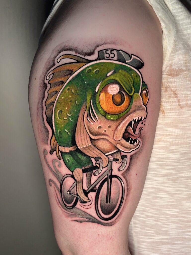 A colorful and quirky tattoo by Sabbe from T-paint/Roxy of a stylized green fish with large orange eyes and sharp teeth. The fish is wearing a hat with the number "55" and is riding a bicycle, with grey exhaust-like swirls behind it.