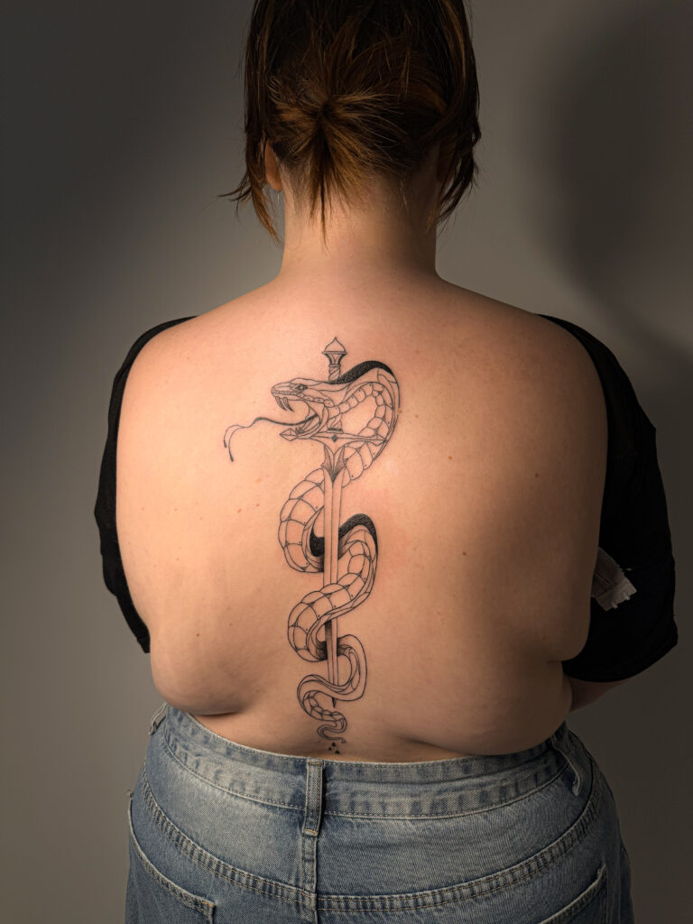 A large, vertical black-ink tattoo on a person's back by Britt of Inked by Britt. It depicts a highly detailed snake with its mouth open, coiled intricately around a long, elegant sword that follows the line of the spine.