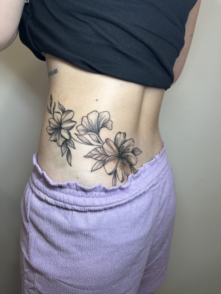 A soft and feminine black and grey floral piece on a client's side/hip area, showcasing the fine shading and botanical style of Laura from Inked By Laura.
