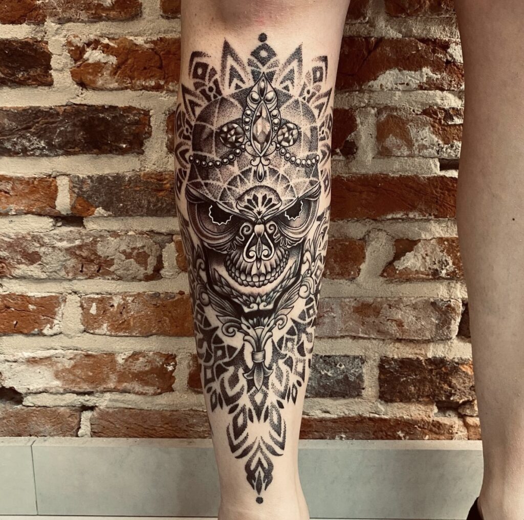 A large, intricate black-ink tattoo on a lower leg by Christa of Obscuria Gratia. This highly detailed piece features an ornamental skull-like figure adorned with crown-like jewels and complex geometric dotwork patterns.