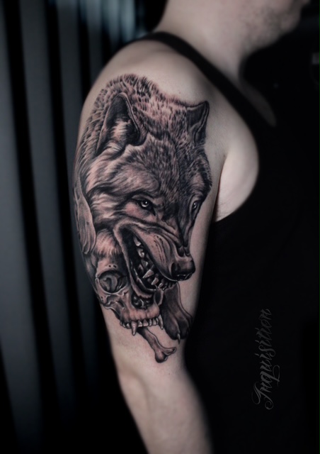 A high-contrast black and grey realism tattoo on an upper arm by Gianni of Inquisition Tattoo. The design features a snarling wolf's head positioned above a human skull that has sharp canine teeth.