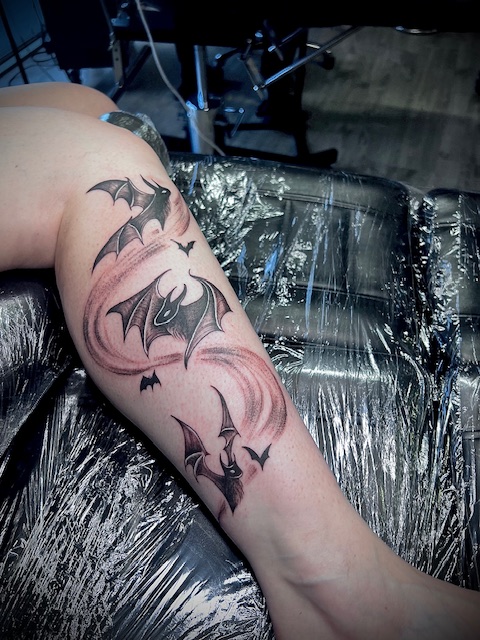 An illustrative black and grey tattoo on a lower leg featuring several bats in flight, surrounded by a swirling trail of shaded energy to create a sense of motion, by Bieke of Bibi Ink.
