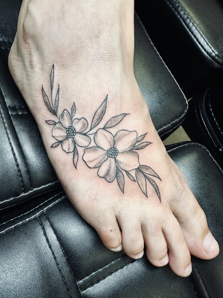 A clean black and grey tattoo of two blooming flowers with leaves on the top of a person's foot, highlighting the precise linework of Laura of Inked By Laura.