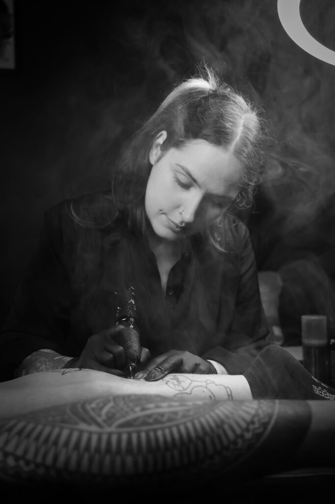 A moody black and white portrait of tattoo artist Christa from Obscuria Gratia. She is deeply focused, leaning over a client’s limb to apply a tattoo while wisps of smoke or mist swirl artistically in the foreground.