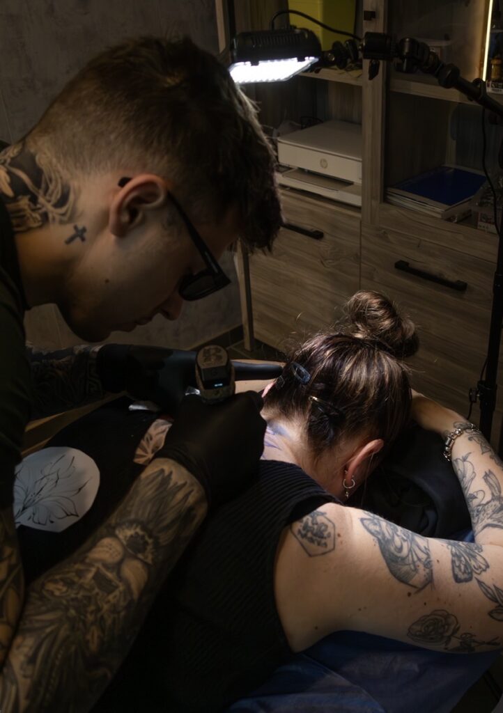 A black and white photograph of tattoo artist Remi from Relife Tattoo focused on his craft. He is wearing glasses and has visible tattoos on his neck and arms while carefully applying a design to a client's upper back.