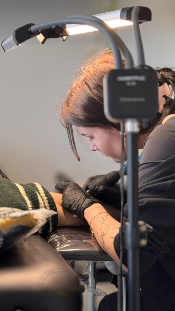 A profile shot of tattoo artist Britt from Inked by Britt focused on her work. She is wearing glasses and black tattooing gloves, leaning over to carefully apply a design to a client's leg under a bright studio light.