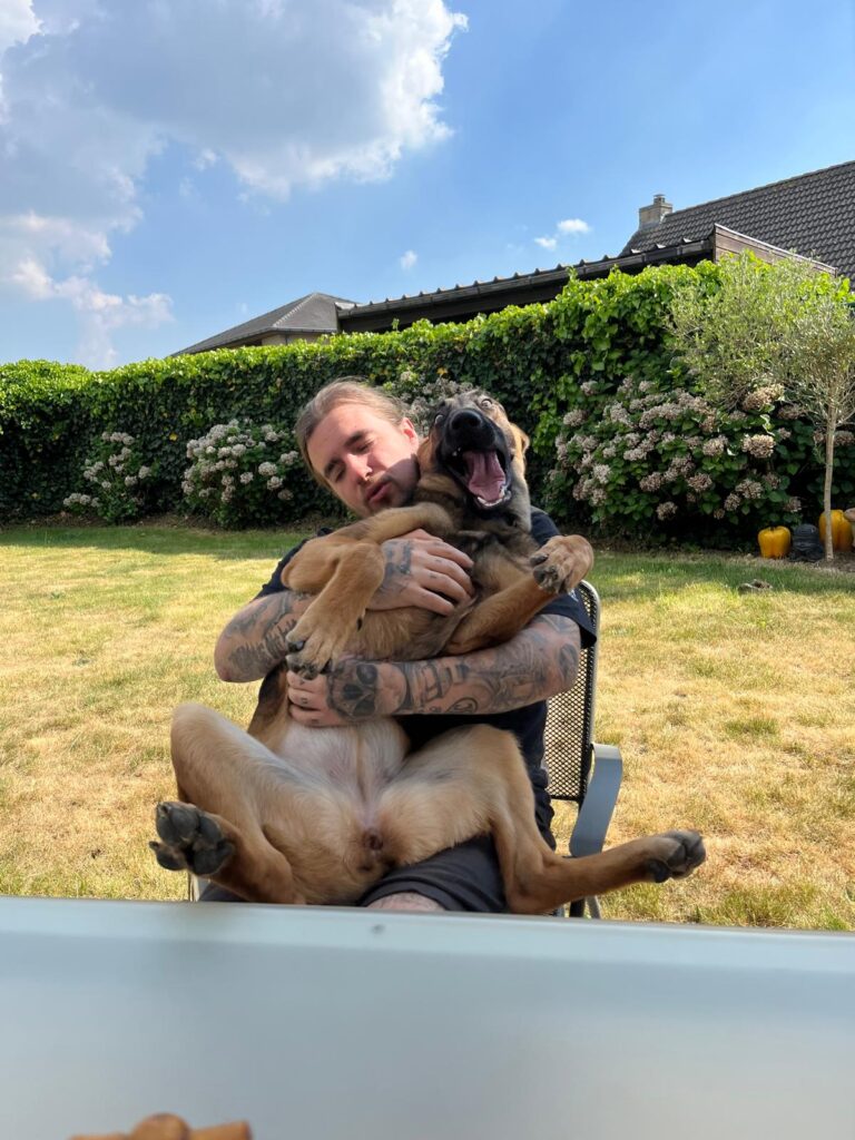 A candid outdoor photo of tattoo artist Gianni from Inquisition Tattoo sitting in a chair and laughing while hugging a large, yawning German Shepherd dog. He has long hair tied back, and his arms are covered in detailed tattoos.