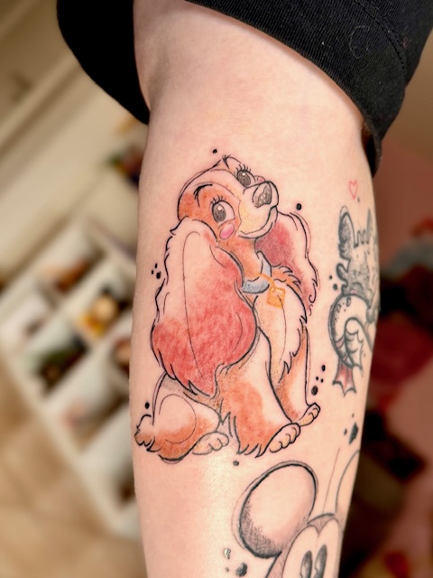 A detailed, illustrative color tattoo of Lady from the Disney classic Lady and the Tramp. The design, by Amber of Fantasy Ink, uses warm tones and soft shading to bring the character to life on an arm.
