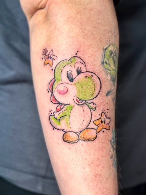 A vibrant tattoo on a forearm depicting the character Yoshi from the Super Mario series, accompanied by two small, cheerful stars. This colorful, pop-culture piece highlights the signature style of Amber from Fantasy Ink.