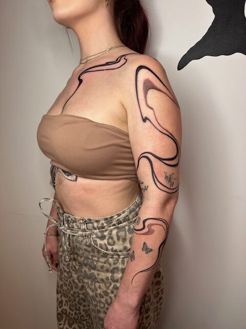 A striking piece by Ezra from TattooGast featuring organic, wave-like black lines that follow the contours of a client's shoulder, upper arm, and chest. The abstract design creates a sense of natural movement across the skin.