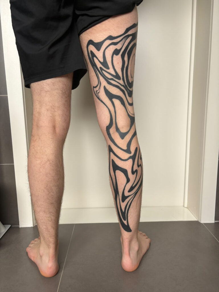 A massive, high-contrast blackwork tattoo on a client's leg by Ezra of TattooGast. The design features thick, solid-black fluid shapes that wrap dynamically around the thigh and calf, weaving between existing tattoos.