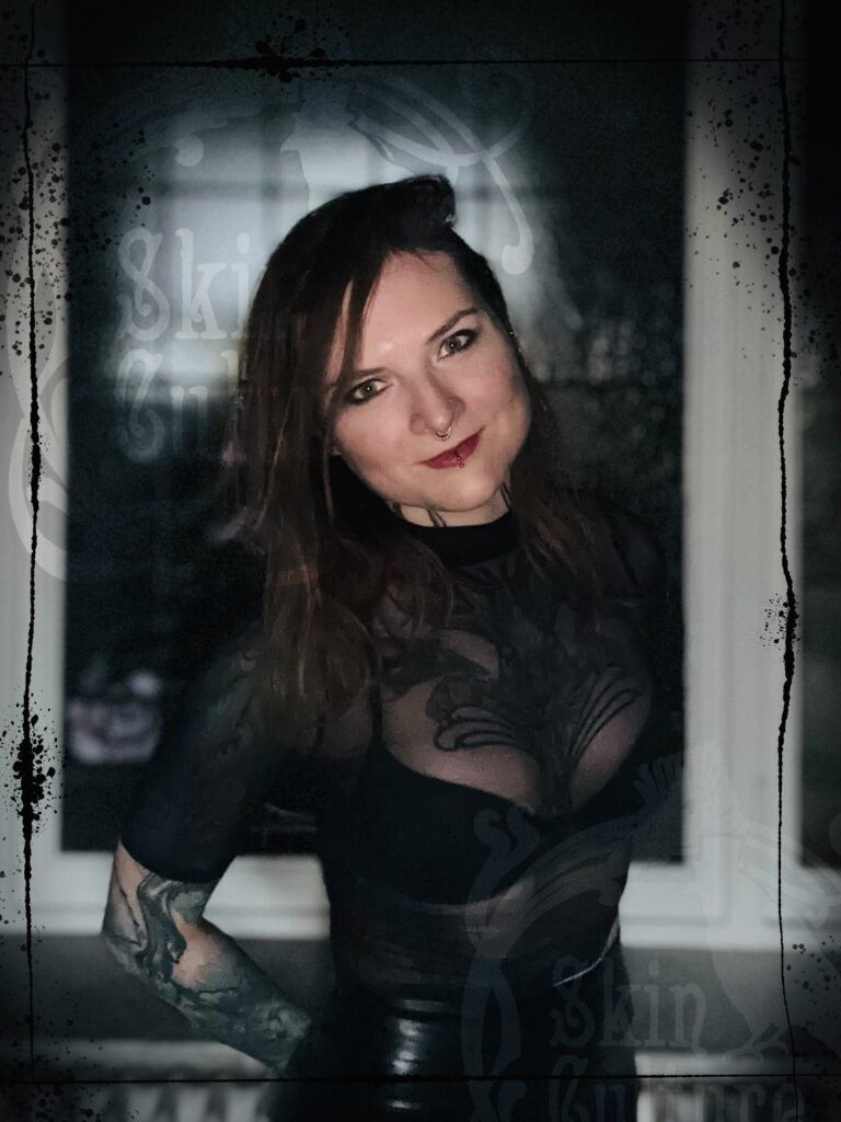 A portrait of tattoo artist Veerle from Skin Culture. She has long, wavy dark-red hair, a septum piercing, and a vertical labret piercing. She is wearing a black sheer top that showcases a large, intricate black-ink mandala and floral tattoo covering her chest.