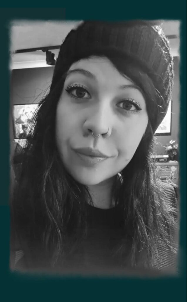 A black and white portrait of tattoo artist Freyja from Deadly Muse. She is wearing a dark beanie and has long hair, with a septum piercing and a vertical labret piercing visible as she looks toward the camera.