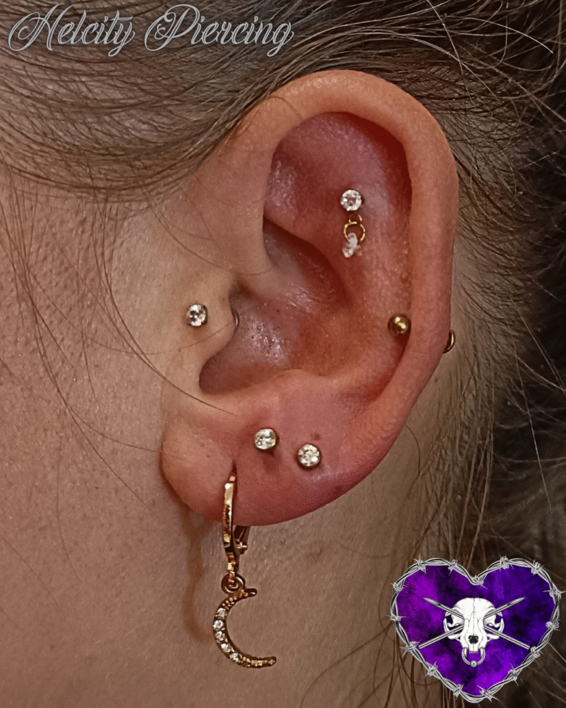 A close-up of an ear featuring a curated collection of gold and jeweled piercings by Milanda from Helcity Piercing. The set includes two stacked lobe studs, a gold crescent moon charm, a tragus piercing, and several helix piercings with delicate dangling details.
