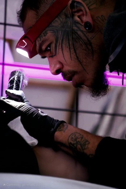 Een sfeervolle close-up van tattoo-artiest Francois (fran6_tatoueur) tijdens de Sint-Niklaas Tattoo Expo. Hij draagt een hoofdlamp en zwarte handschoenen, terwijl hij geconcentreerd een tatoeage zet. Verschillende tattoos zijn zichtbaar op zijn gezicht, nek en handen.