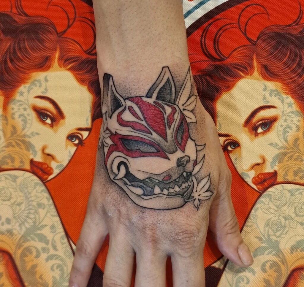 Een kleurrijke tattoo op een handrug van een Japans Kitsune (vos) masker, uitgevoerd in een gedetailleerde stijl met rode en grijze tinten. Het masker is omringd door bladeren en de achtergrond toont een artistiek patroon van een getatoeëerde vrouw op een rode ondergrond. Werk van Francois (fran6_tatoueur) op de Sint-Niklaas Tattoo Expo.