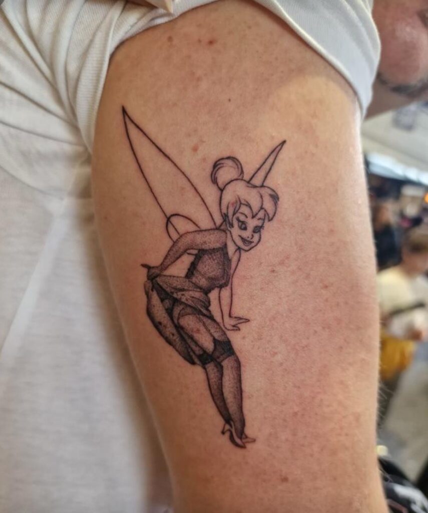 Een speelse black and grey tattoo op een bovenarm van Tinkerbell in een pin-up pose. Ze tilt haar rokje op en draagt jarretels, uitgevoerd met fijn lijnwerk en dotwork-schaduwen. Gezet door Francois van fran6_tatoueur tijdens de Sint-Niklaas Tattoo Expo.