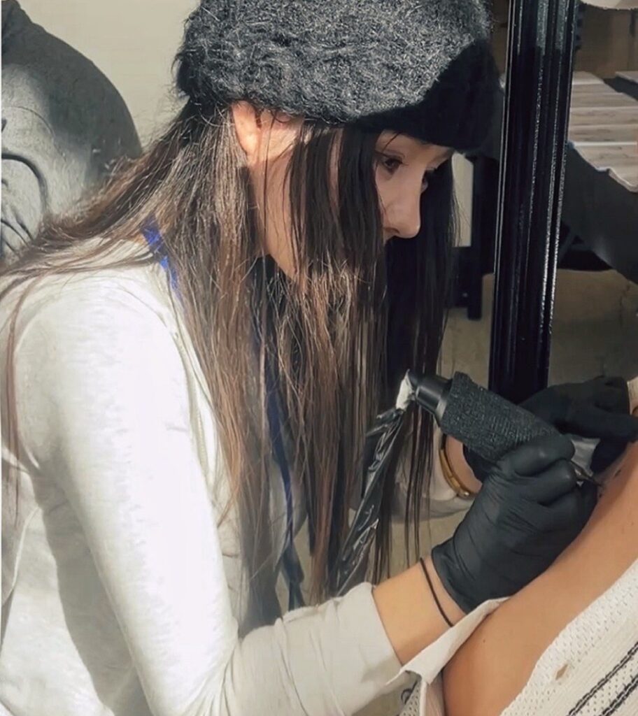 Een sfeervolle actiefoto van tattoo-artiest Yifei (Herz Tattoo) op de Sint-Niklaas Tattoo Expo. Ze draagt een zwarte muts en zwarte handschoenen en werkt met uiterste precisie aan een tatoeage op een arm. Haar focus op het fijne detailwerk is duidelijk zichtbaar.