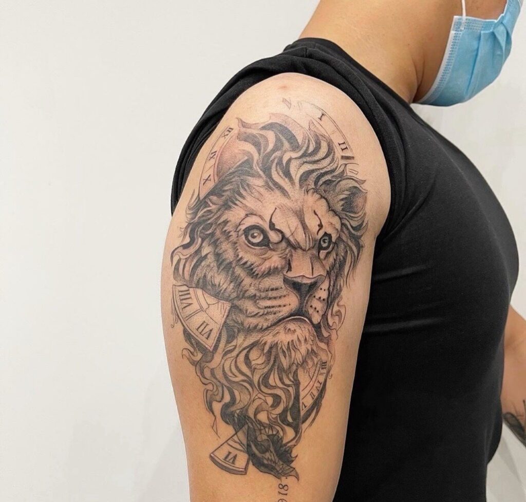Een krachtige black and grey tattoo op een bovenarm van een leeuwenkop. Het ontwerp is artistiek gecombineerd met fragmenten van een klok en Romeinse cijfers, uitgevoerd met zachte schaduwen en strakke lijnen. Gezet door Yifei van Herz Tattoo op de Sint-Niklaas Tattoo Expo.